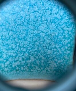 10x magnification of my strontium matt turquoise glaze looking through a hand lens. You can see the crystals on the surface. The turquoise glaze is underneath and the paler matt crystals have grown on the surface. This phase separation causes a mottled effect. This glaze contains copper oxide and titanium dioxide.