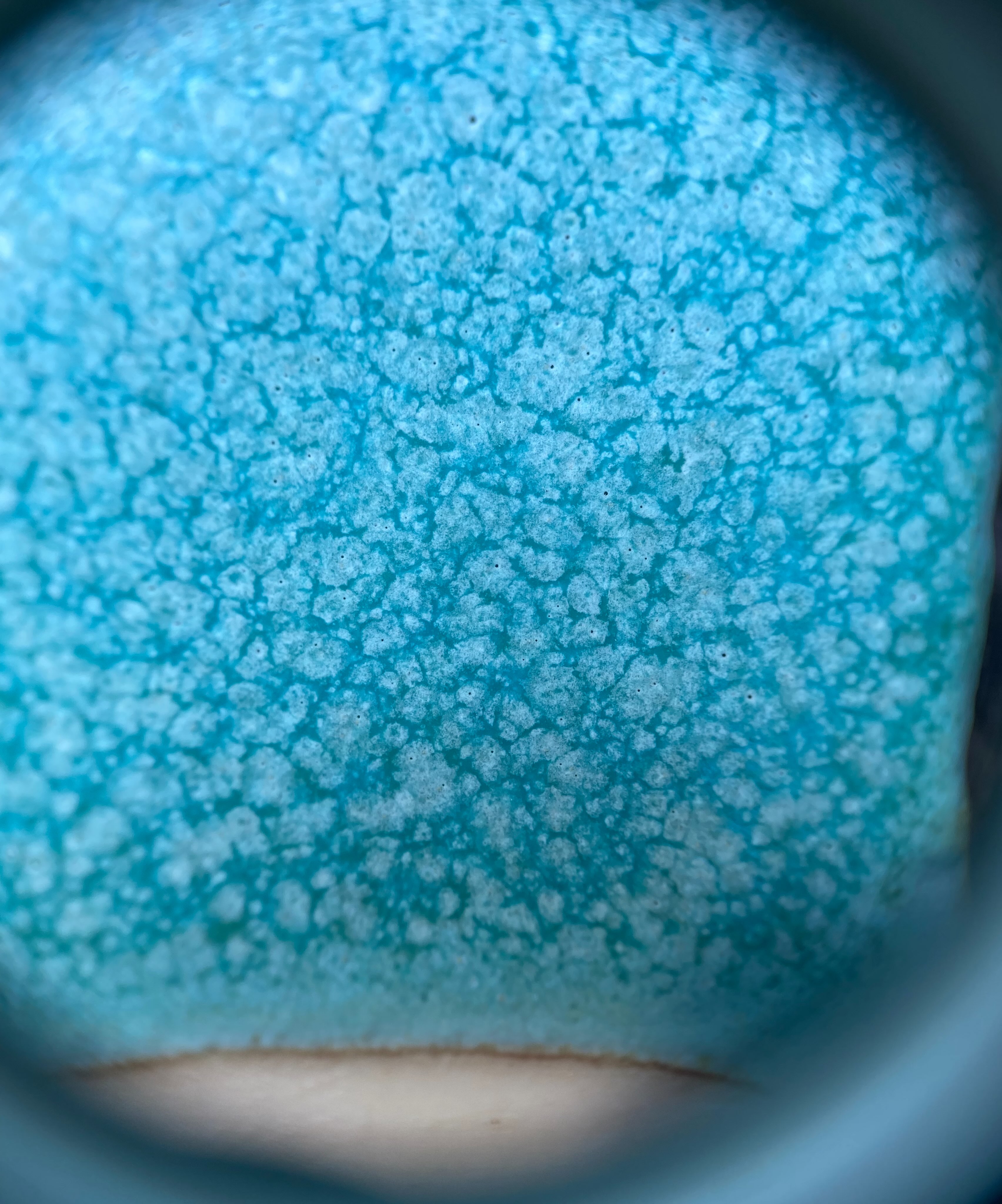 10x magnification of my strontium matt turquoise glaze looking through a hand lens. You can see the crystals on the surface. The turquoise glaze is underneath and the paler matt crystals have grown on the surface. This phase separation causes a mottled effect. This glaze contains copper oxide and titanium dioxide.